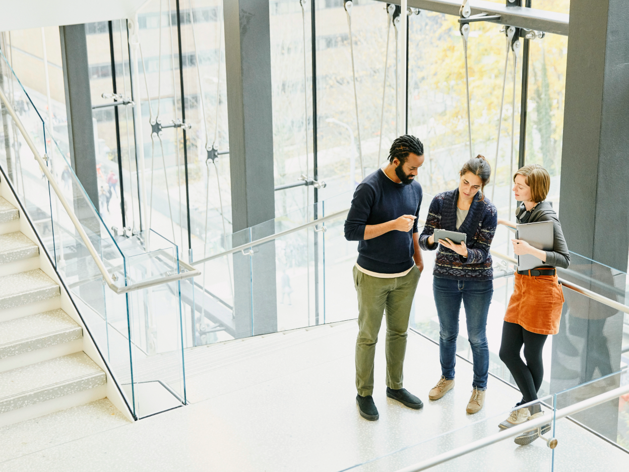 Landscape 5500 pixel wide by 3667 pixel high jpg photo of three employees in their 20s holding a conversation on a set of stairs in an office building's lobby. Large windows can be seen in the background.